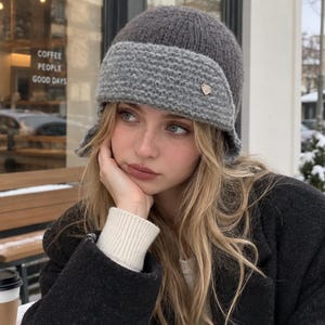 May include: A two-toned knit beanie featuring a dark charcoal gray crown and a lighter gray textured brim. The hat includes a small, metallic, triangular-shaped charm attached to the side of the brim, adding a subtle detail to the knit construction.