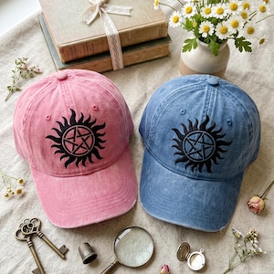 May include: Two baseball caps, one pink and one blue, each with a black sun and pentagram design. The caps are on a light-colored surface with vintage books, flowers, and antique accessories.