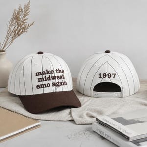 May include: Two white baseball caps with thin black pinstripes and brown brims. The front cap features the embroidered brown text "make the midwest emo again". The back cap displays the embroidered brown number "1997" above the adjustable strap.