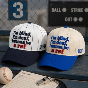May include: Two baseball caps with pinstripes and embroidered text reading I'm blind, I'm deaf, I wanna be a ref. One cap is white with a navy brim and RFD lettering, the other is cream with a blue brim and BLF lettering, both featuring top buttons.