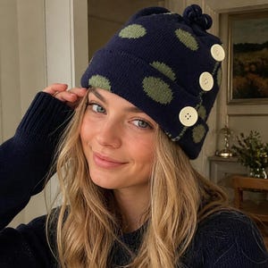 May include: A navy blue knit beanie featuring a pattern of olive green polka dots. The hat has a folded cuff adorned with three large, round, cream-colored buttons arranged vertically along the side and a small knit knot detail at the top.