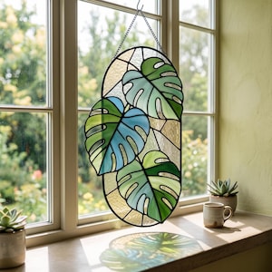 May include: An oval stained glass suncatcher featuring three monstera leaves in shades of blue and green, framed in black metal. The piece is suspended by a thin metal chain and displays a textured, translucent background with clear glass segments.