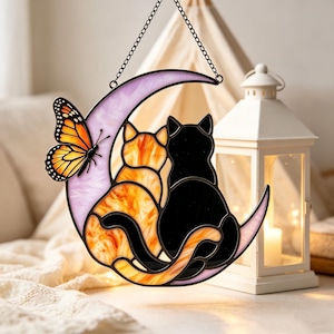 May include: A stained glass suncatcher featuring a crescent moon in light purple, an orange and black monarch butterfly, and two cats, one orange tabby and one solid black, sitting together on the moon, suspended by a black metal chain.