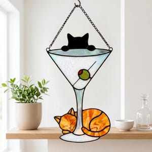 May include: A stained glass suncatcher featuring a martini glass with a green olive, a black cat silhouette peering over the rim, and an orange tabby cat curled around the base, all suspended by a black metal chain for hanging display.