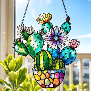 May include: A stained glass suncatcher featuring a collection of green cacti in a multicolored mosaic pot. The cacti are adorned with vibrant yellow, white, purple, and pink flowers. The piece is suspended by a black metal chain for hanging.