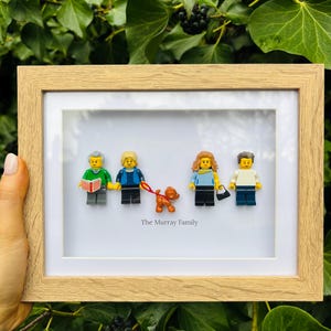 May include: A framed Lego family portrait with a dog, set in a wooden frame with a white mount. The text "The Murray Family" is below the figures. The background is lush green foliage. A unique and personalised gift idea.