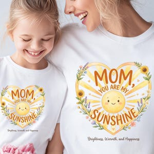 May include: White t-shirts with a heart-shaped design featuring a smiling sun, sunflowers, and the text "MOM, YOU ARE MY SUNSHINE." The shirts also include the text "Brightness, Warmth, and Happiness."