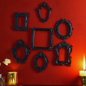 Może przedstawiać: Kolekcja siedmiu ozdobnych, czarnych ramek do zdjęć o różnych kształtach, w tym kwadratowych, owalnych i łukowych, jest ułożona na żywej czerwonej ścianie. Ramki mają szczegółowe, dekoracyjne krawędzie. Zapalona świeca i dekoracyjne szkło znajdują się na pierwszym planie.