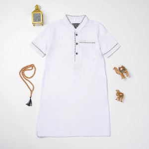 May include: White short-sleeved tunic with a mandarin collar and a pocket. The tunic has decorative trim and buttons. A prayer bead necklace, a gold lantern, and camel figurines are also in the image.