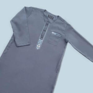 May include: A gray tunic with long sleeves and a mandarin collar. The tunic features a decorative embroidered panel down the front, a pocket, and three buttons. The label on the collar reads "ZAINAB MUSLIM".