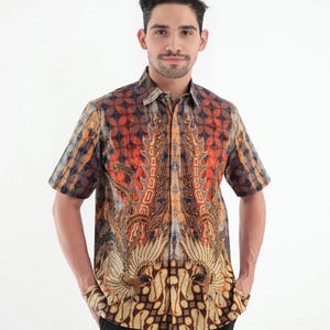 Puede incluir: Camisa de manga corta con estampado batik en tonos naranja, marrón y azul. La camisa presenta un diseño detallado con motivos simétricos y un cuello clásico. El modelo lleva pantalones negros.