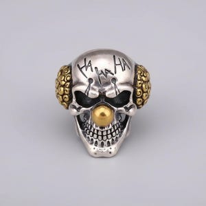 Puede incluir: Anillo de calavera plateado con diseño de payaso. El anillo presenta una nariz y detalles en las orejas de color dorado, con la palabra "HA HA HA" grabada en la frente. La calavera tiene una sonrisa detallada con la boca abierta.