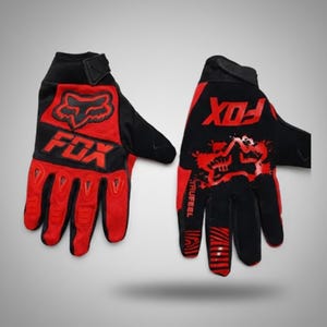 Puede incluir: Un par de guantes de carreras Fox negros y rojos. Los guantes presentan el logotipo de Fox en rojo en el dorso de la mano y la palabra "FOX" en rojo. Los guantes tienen una correa de mu&ntilde;eca negra y detalles rojos en los dedos.