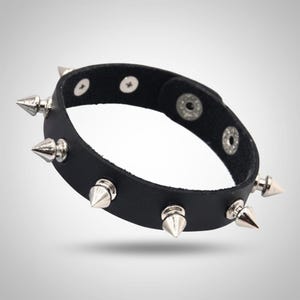 Puede incluir: Pulsera de cuero negro con tachuelas cónicas plateadas. La pulsera tiene un cierre de botón a presión. Las tachuelas están espaciadas uniformemente alrededor de la banda, creando una estética punk rock o alternativa.