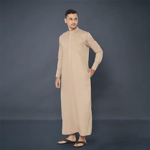 Op de afbeelding: Een beige thobe met lange mouwen, een opstaande kraag en een enkele borstzak. Het kledingstuk is vloerlang en wordt gedragen met donkerbruine sandalen. De achtergrond is effen, donkergrijs.