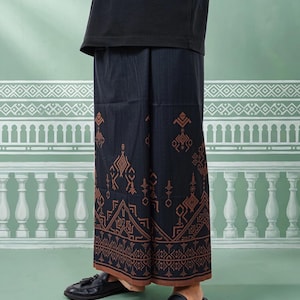 May include: A black sarong with a brown geometric pattern. The sarong is worn with a black t-shirt and black loafers. The background is a light green color with a white decorative design.