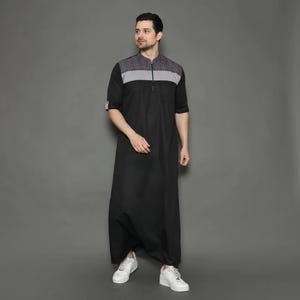 May include: A black thobe with a gray and checkered panel across the shoulders and chest. The thobe has a zipper closure and short sleeves. The person in the image is wearing white sneakers.