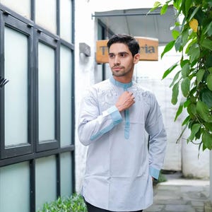 May include: A light gray tunic with intricate embroidery on the chest and light blue accents on the collar and cuffs. The long-sleeved shirt is paired with black pants. The tunic has a mandarin collar.