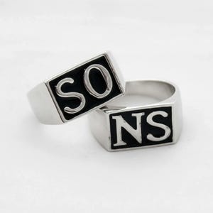 May include: Two silver rings with black rectangular insets. One ring displays the letters "SO" in white, and the other displays "NS". The rings have a polished finish and a classic signet ring design.