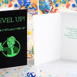 Op de afbeelding: Zwarte wenskaart met de groene tekst "LEVEL UP!". De kaart toont een stripfiguur die een duim omhoog steekt en de tekst "+1 year | +10 Luck | +100 XP". De binnenkant van de kaart heeft een groene boodschap.