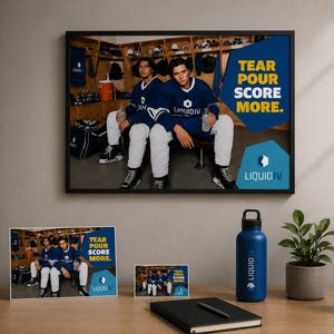 Op de afbeelding: Een ingelijste wandposter en twee kleinere prints met twee hockeyspelers in blauwe shirts in een kleedkamer met de tekst TEAR POUR SCORE MORE en LIQUID IV. Een blauwe waterfles met het LIQUID IV logo staat op een houten bureau.