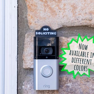 May include: A silver and black video doorbell mounted on a stone wall with a black sign attached to the top that reads NO SOLICITING in white text. A green-bordered starburst graphic on the right side contains the text NOW AVAILABLE IN DIFFERENT COLORS.