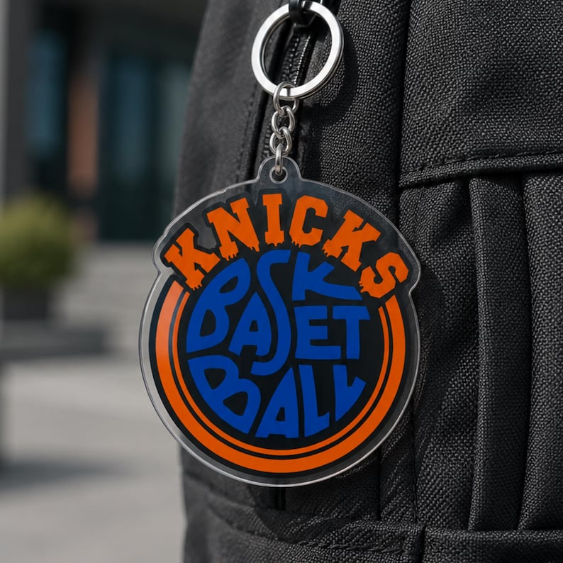 Pu&ograve; includere: Un portachiavi in acrilico trasparente con le parole "Knicks Basketball" in arancione e blu. Il portachiavi &egrave; attaccato a un portachiavi e una catena argentati. Il portachiavi &egrave; appeso a uno zaino nero.