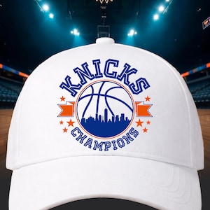Pu&ograve; includere: Cappellino da baseball bianco con la scritta "Knicks Champions" in blu, un pallone da basket e un design di skyline. Il cappellino ha nastri arancioni e stelle, per gli appassionati di sport.