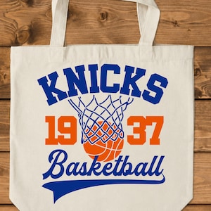 Pu&ograve; includere: Borsa tote color crema con la scritta "KNICKS" in blu, una grafica di rete e palla da basket. I numeri "1937" sono arancioni e la parola "Basketball" &egrave; blu.
