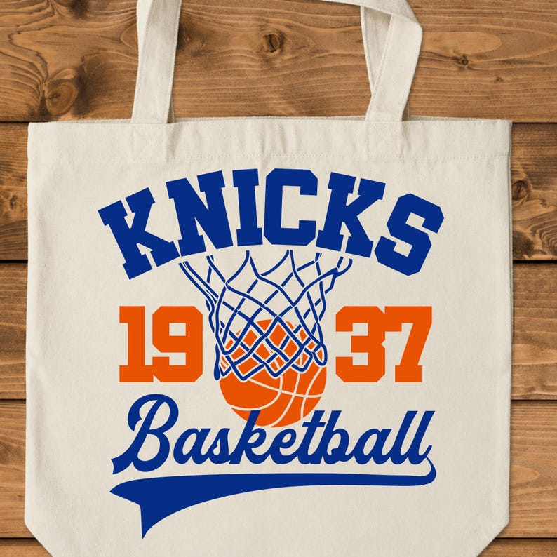 Pu&ograve; includere: Borsa tote color crema con la scritta "KNICKS" in blu, una grafica di rete e palla da basket. I numeri "1937" sono arancioni e la parola "Basketball" &egrave; blu.
