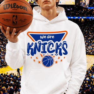 Pu&ograve; includere: Felpa con cappuccio bianca con la scritta "we are KNICKS" in blu e arancione, insieme a una grafica di basket. Nell'immagine &egrave; tenuto un pallone da basket marrone con il logo Wilson.