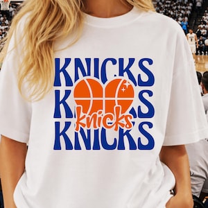 Pu&ograve; includere: T-shirt bianca con la scritta "Knicks" in blu e una grafica di pallone da basket arancione. La parola "Knicks" &egrave; ripetuta tre volte, con la grafica del pallone da basket al centro. La maglietta &egrave; in stile casual.