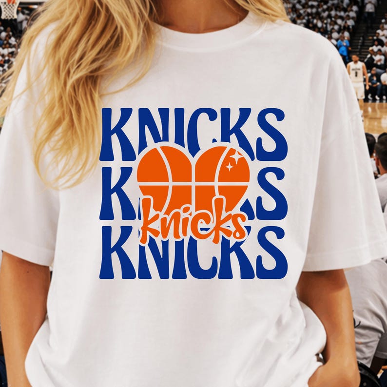 Pu&ograve; includere: T-shirt bianca con la scritta "Knicks" in blu e una grafica di pallone da basket arancione. La parola "Knicks" &egrave; ripetuta tre volte, con la grafica del pallone da basket al centro. La maglietta &egrave; in stile casual.