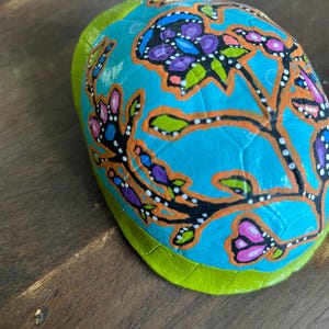 May include: A hand-painted, oval-shaped decorative item with a bright turquoise background. The item features a floral design with purple, pink, and orange accents. The edges are outlined in green. The item is placed on a wooden surface.