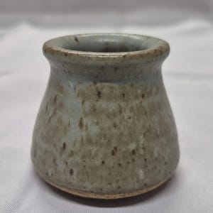 以下が含まれることがあります： 広い口と広がった縁を持つ小さな陶器のポット。外側はライトグレー、ベージュ、ブラウンの色合いの斑点のある釉薬で仕上げられており、滑らかで丸みを帯びた本体は底に向かって細くなっており、釉薬のかかっていない素焼きの土が見えています。