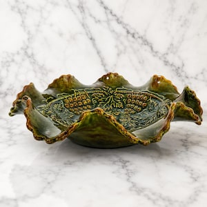 May include: A round, glazed ceramic bowl in a deep olive green hue, featuring a decorative embossed pattern of grape clusters and vine leaves. The bowl has a distinctively wavy, ruffled edge that creates a scalloped silhouette around the rim.