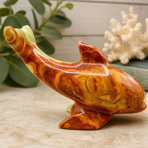 May include: A handcrafted dolphin figurine with a polished finish. The sculpture features a blend of orange, brown, and yellow hues, with green accents on the fins. The dolphin is posed in a leaping position, showcasing its streamlined form.