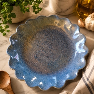 May include: A round ceramic plate with a scalloped edge, featuring a speckled glaze in shades of blue and hints of golden brown. The surface has a textured, mottled appearance with a glossy finish, creating a distinct color gradient across the center.