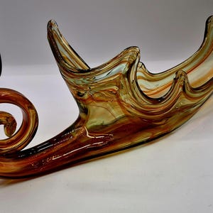 May include: A decorative glass bowl with a swirling design in shades of amber, brown, and orange. The bowl has a unique shape with a curled end and undulating edges, perfect for home decor.
