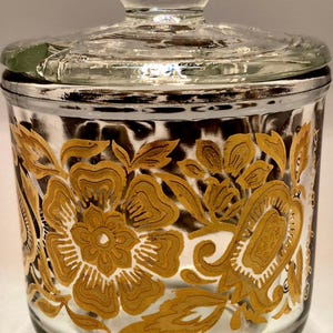 May include: A clear glass jar with a lid, featuring a gold floral design. The jar has a silver rim and a clear knob on the lid. The floral pattern is detailed and covers the jar's surface.