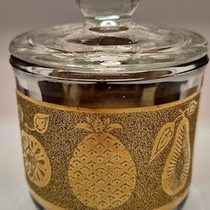 May include: A clear glass jar with a lid, featuring a gold band with fruit designs. The jar has a pineapple, pear, and other fruit illustrations. The lid has a small knob for easy opening. The jar is likely for storing food items.