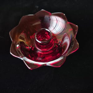May include: A ruby red glass candle holder with a flower-shaped base. The candle holder has a central cylindrical cup for the candle and a scalloped edge. The glass is transparent, allowing light to pass through, and reflects light.