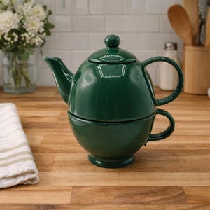 Könnte beinhalten: Ein dunkelgrünes Keramik-Teeservice, bestehend aus einer Teekanne und einer Tasse. Die Teekanne hat eine runde Form mit Deckel und Ausguss. Die Tasse ist unter der Teekanne gestapelt. Das Set steht auf einer Holzoberfläche.