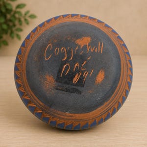 May include: A circular ceramic base with a dark grey centre and a blue outer rim featuring a carved leaf pattern. The centre is inscribed with the handwritten text Coggeshall and the date 09 in orange, with a small orange smudge near the edge.