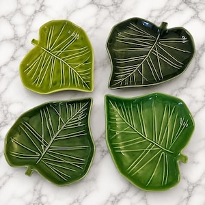 May include: A set of four ceramic dishes shaped like leaves, each featuring a glossy finish and raised white vein details. The collection includes one light green dish, one dark forest green dish, and two medium green dishes with distinct textures.