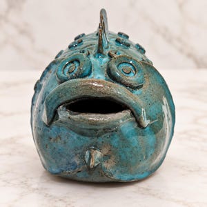Puede incluir: Una escultura de pez de cerámica en color verde azulado con acabado brillante, que presenta ojos circulares prominentes, una boca ancha abierta y detalles texturizados en relieve a lo largo de la cresta superior. La superficie muestra variaciones en tonos azules y verdes con acentos marrones.
