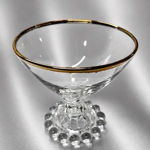 Pode incluir: Uma taça de sobremesa em vidro transparente com uma abertura larga e um rebordo dourado metálico. A taça assenta numa base curta e canelada, rodeada na parte inferior por um anel de pequenas esferas de vidro transparente decorativas.