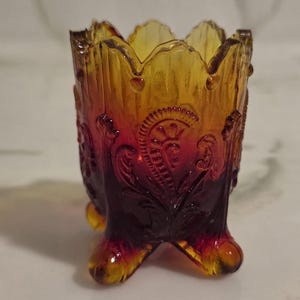 Puede incluir: Un pequeño palillero de vidrio con patas que presenta un degradado vibrante, pasando de un rojo oscuro profundo en la base a un ámbar translúcido en el borde festoneado, decorado con un patrón floral en relieve tipo cachemir.