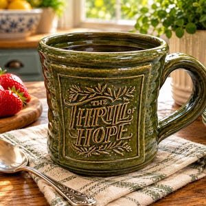 May include: A dark green ceramic mug with a glossy finish and a textured, ribbed exterior. The front features a raised, rectangular panel with the embossed text "THE THRILL OF HOPE" surrounded by decorative leaf motifs and a sturdy curved handle.