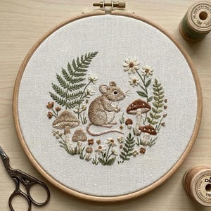 Puede incluir: Obra bordada con un ratón marrón rodeado de setas, helechos y flores, en un aro de madera. También se ven dos carretes de hilo y unas tijeras. Paleta de colores naturales y bordado detallado.